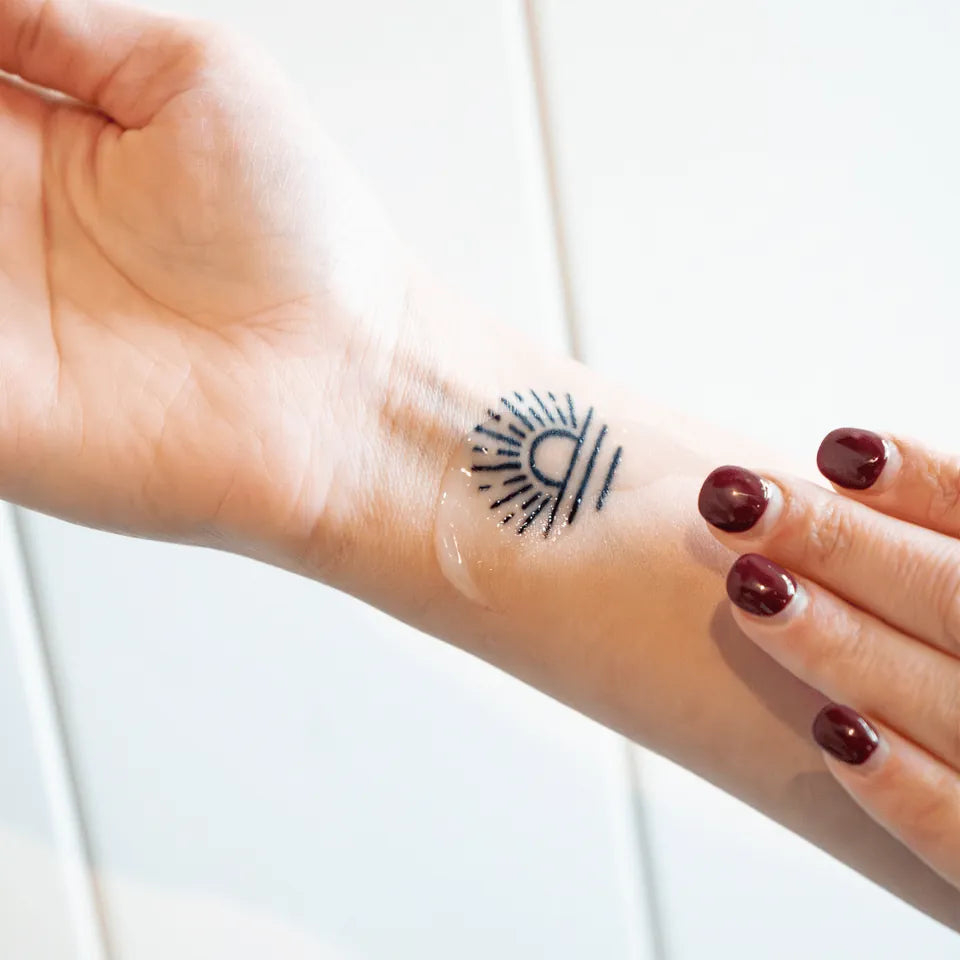 A hand with dark red nail polish applies Amaroo Naturals emu oil to a sun tattoo on a wrist